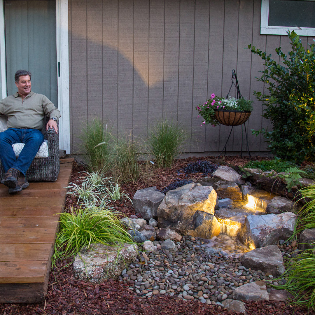 Man sitting on a chair by a small garden pond with stones and decorative ground cover